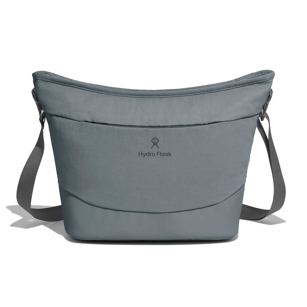 Person in olive sweater and dark jeans holding gray Hydro Flask insulated tote by a black shoulder strap. Puffy upper, reinforced bottom panel, and logo on front.